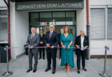 Pred vhodom v zdravstveni dom stojijo dva moška in dve ženski. V rokah imajo škarje in se pripravljajo na rezanje traku v barvah slovenske zastave. Za njimi stojita še dva moška.
