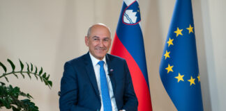 Janez Janša sedi, v ozadju na levi je rastlina, na desni so zastave.